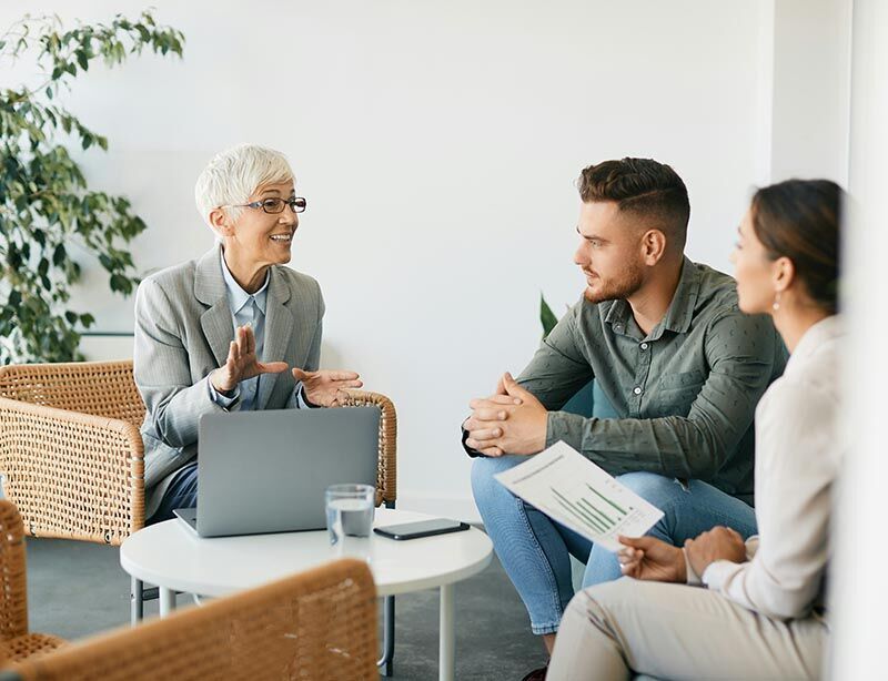 Eine Beraterin sitzt mit ihrem aufgeklappten Laptop zusammen mit ihren Kunden, einem Mann und einer Frau, um einen runden Tisch und erklärt etwas.